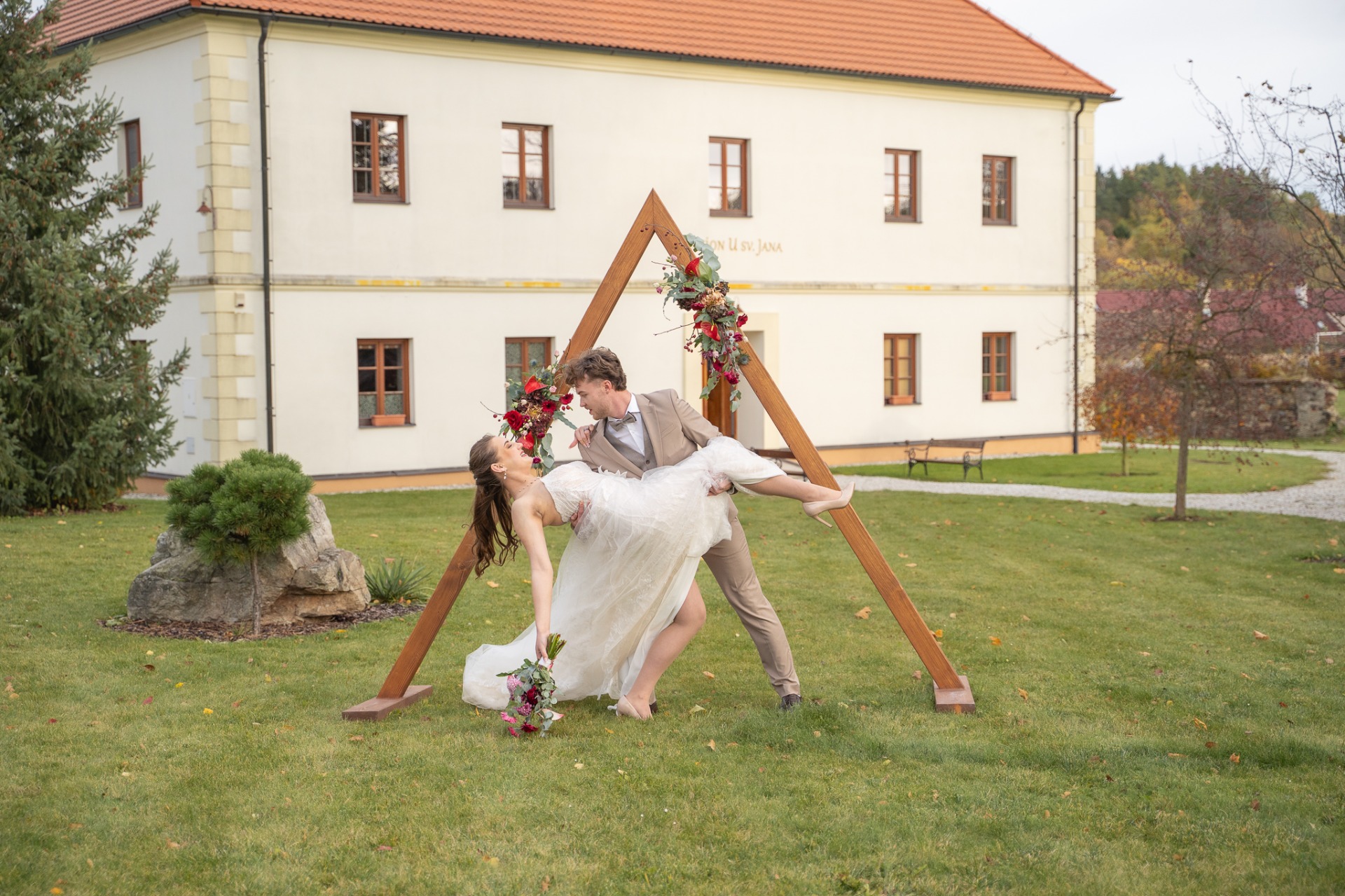 Nevěsta a ženich na trávníku před dřevěnou slavobránou s květinami před bílou budovou penzionu